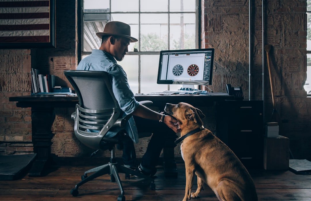 A man in a hat sits at a desk in a casual office, playfully interacting with a dog. A computer screen displays graphics, capturing a lighthearted moment.