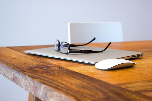 A closed laptop with glasses on top and a wireless mouse on a wooden desk, suggesting a setup for a subtle workplace prank.