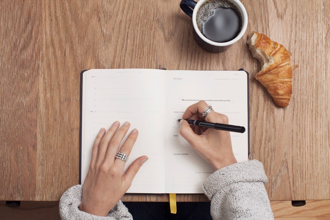 Person writing in a planner at a desk with coffee and a croissant, suggesting planning an office prank to boost team morale.